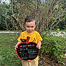 Tatum joined the competition — help win amazing prizes! child, boy, outdoor, grass, bush, tree, sign, yellow_shirt, shorts, first_day, pre_k, person, portrait, holding, smiling, nature, greenery, daytime, cute, young