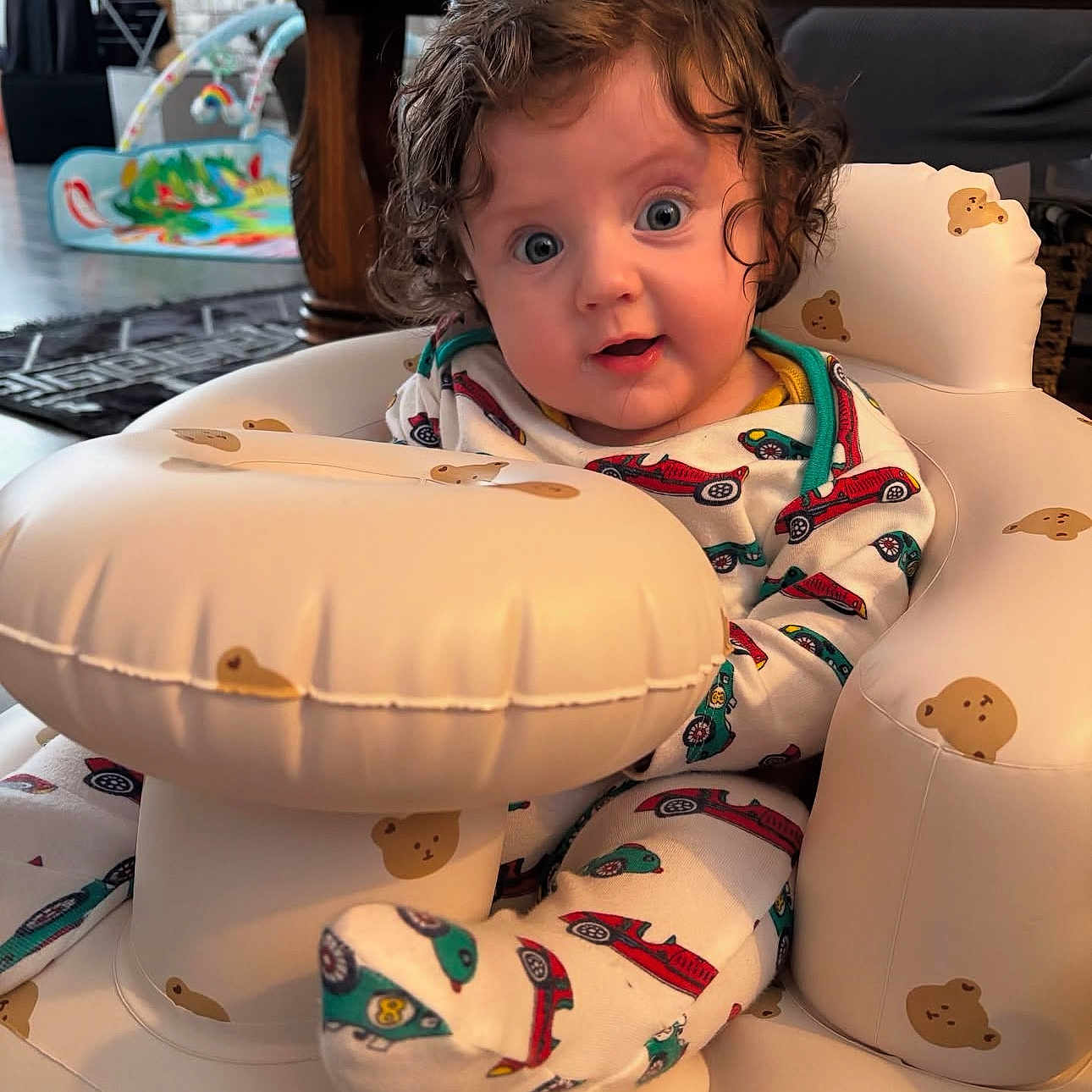 Evan joined the competition — help win amazing prizes! baby, baby_chair, bright_colors, child, colorful_toys, curly_hair, cushion, cute, expression, face, floor_rug, footwear, indoor, infant, onesie, person, playroom, seat, teddy_bear_pattern, wooden_table