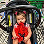 baby, child, car_seat, shopping_cart, red_dress, hair_bows, girl, fabric, store, retail, product_shelves, floor, plastic_cup, seat_belt, person, indoor, shopping, cute, infant, chewing