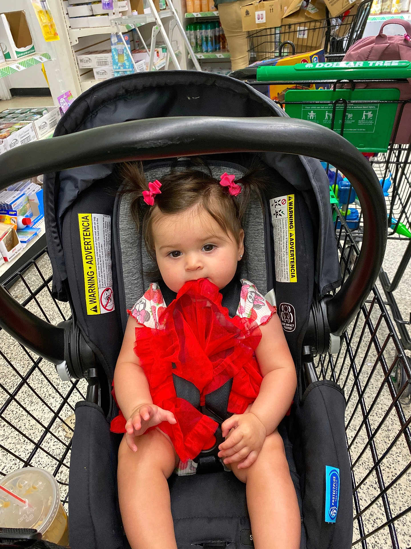 Vayda is registered to the contest to win money with this photo: baby, child, car_seat, shopping_cart, red_dress, hair_bows, girl, fabric, store, retail, product_shelves, floor, plastic_cup, seat_belt, person, indoor, shopping, cute, infant, chewing