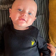 Reggie is registered to the contest to win money with this photo: baby, infant, face, blue_eyes, black_clothing, yellow_logo, hand, blanket, striped_fabric, lying_down, cute, portrait, child, skin, head, smile, human, young, tender, cozy