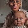 Reggie is registered to the contest to win money with this photo: woman, baby, child, glasses, curly_hair, knit_sweater, indoor, portrait, holding, calm, face, closeup, skin, hand, expression, person, adult, young_child, cozy, warm_lighting