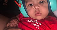 Legend is registered to the contest to win money with this photo: baby, child, headphones, red_clothing, hands, person, indoor, closeup, face, infant, holding, skin, expressionless, noise_cancelling_headphones, dark_background, cute, portrait, young_child, human, headgear