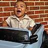 toddler, child, toy_car, smiling, happy, brick_wall, outdoor, short_sleeve_shirt, cute, playful, vehicle, baby, joyful, portrait, person, seat, expression, fun, car, small