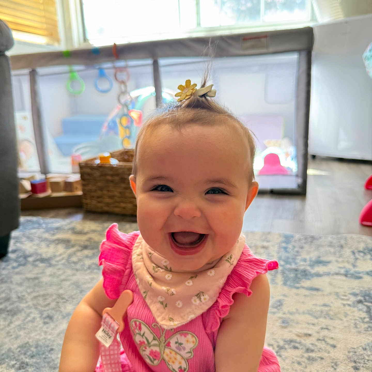 Raylee is registered to the contest to win money with this photo: baby, child, smiling, pink_dress, bib, hair_clip, indoor, playpen, toy, rug, happy, cute, infant, childhood, person, portrait, sitting, daylight, home, floor