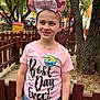 child, birthday, pink_shirt, mickey_ears, headband, outdoor, fence, tree, happy, smile, jeans, phone, badge, park, celebration, daylight, person, casual_clothing, event, nature
