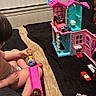 bed, bedroom, black_bedspread, blanket, carpet_strip, child, denim_knee, doll, hat_on_wall, indoor, jacket_on_hook, miniature_furniture, pink_ramp, plastic_toy, playtime, toddler, toy_car, toy_house, toys, window_blinds