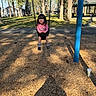 bicycle, blue_pole, boots, child, daytime, fun, grass, house, nature, outdoor, park, person, pink_shirt, playground, shadow, smiling, sunlight, swing, trees, woodchips