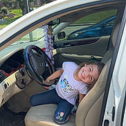 Harmony is registered to the contest to win money with this photo: building, car_interior, child, dashboard, door, floor_mat, greenery, happy, jeans, person, playful, seat, seatbelt, smiling, steering_wheel, sunlight, tshirt, vehicle, white_car, window