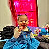child, toddler, blue_dress, denim, indoor, bed, blanket, toy, red_curtain, smile, snack, happy, hair_buns, person, clothing, sitting, room, window, colorful, casual