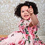 child, toddler, smiling, pointing, curly_hair, floral_dress, pink, wooden_surface, bokeh_background, portrait, happy, cute, sitting, indoor, fashion, person, photography, studio, playful, girl