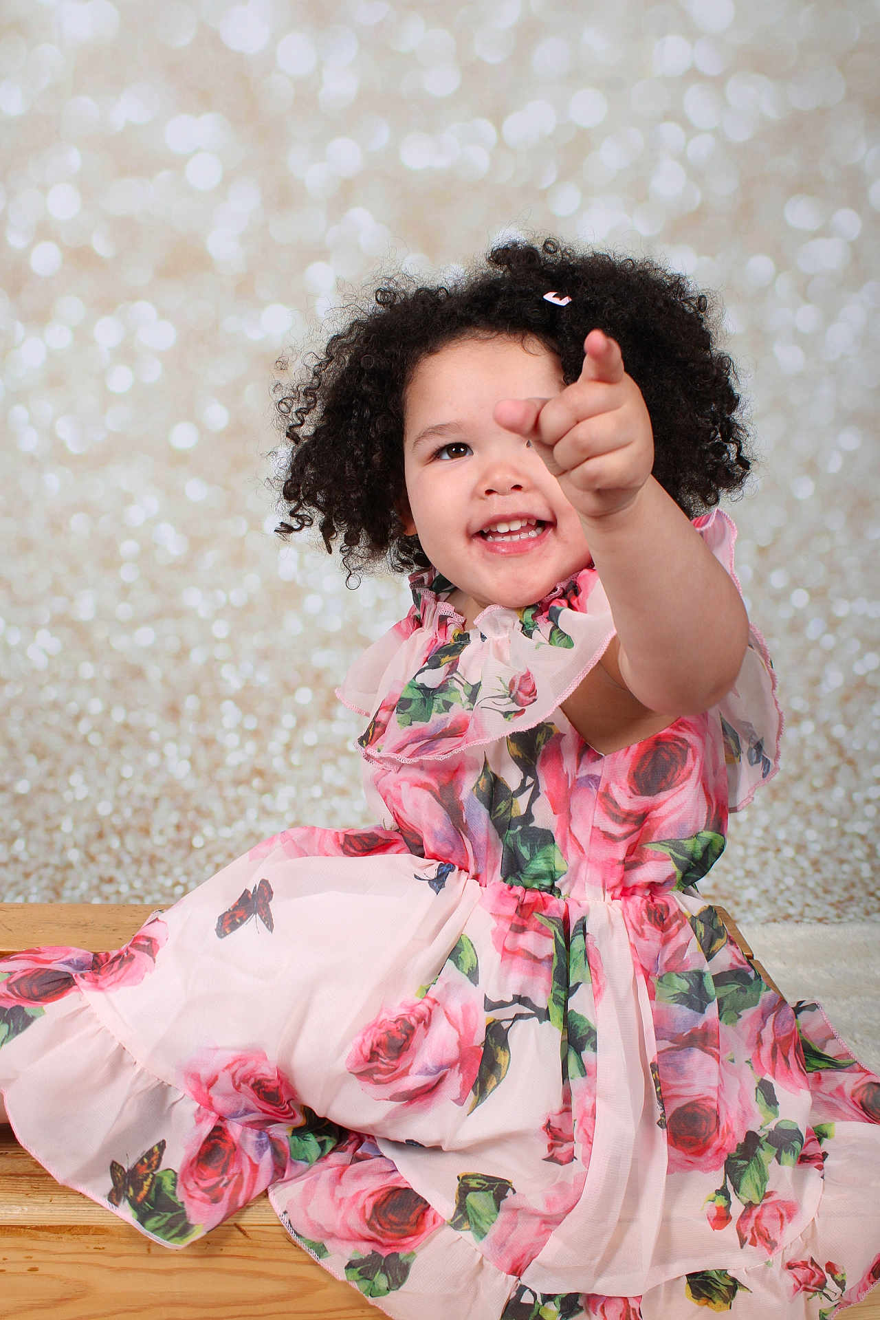 Zendaya joined the competition — help win amazing prizes! child, toddler, smiling, pointing, curly_hair, floral_dress, pink, wooden_surface, bokeh_background, portrait, happy, cute, sitting, indoor, fashion, person, photography, studio, playful, girl