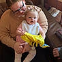 Payton joined the competition — help win amazing prizes! baby, adult, glasses, pink_clothing, bib, plush_toy, yellow_toy, bee_toy, couch, leather_couch, indoor, furniture, wooden_cabinet, figurine, green_frog, carpet, happy, smiling, holding, person