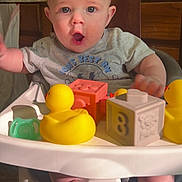 Zander joined the competition — help win amazing prizes! baby, high_chair, rubber_duck, toy_block, foot, expression, indoor, child, play, face, clothing, tabletop, wooden_chair, curious, infant, pacifier, plastic_toy, smile, person, furniture
