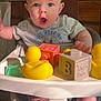 baby, high_chair, rubber_duck, toy_block, foot, expression, indoor, child, play, face, clothing, tabletop, wooden_chair, curious, infant, pacifier, plastic_toy, smile, person, furniture