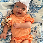 Violet is registered to the contest to win money with this photo: baby, infant, child, orange_outfit, headband, bow, blanket, pattern, blue, white, smiling, tongue_out, cute, happy, lying_down, fabric, clothing, soft_toys, pacifier, person