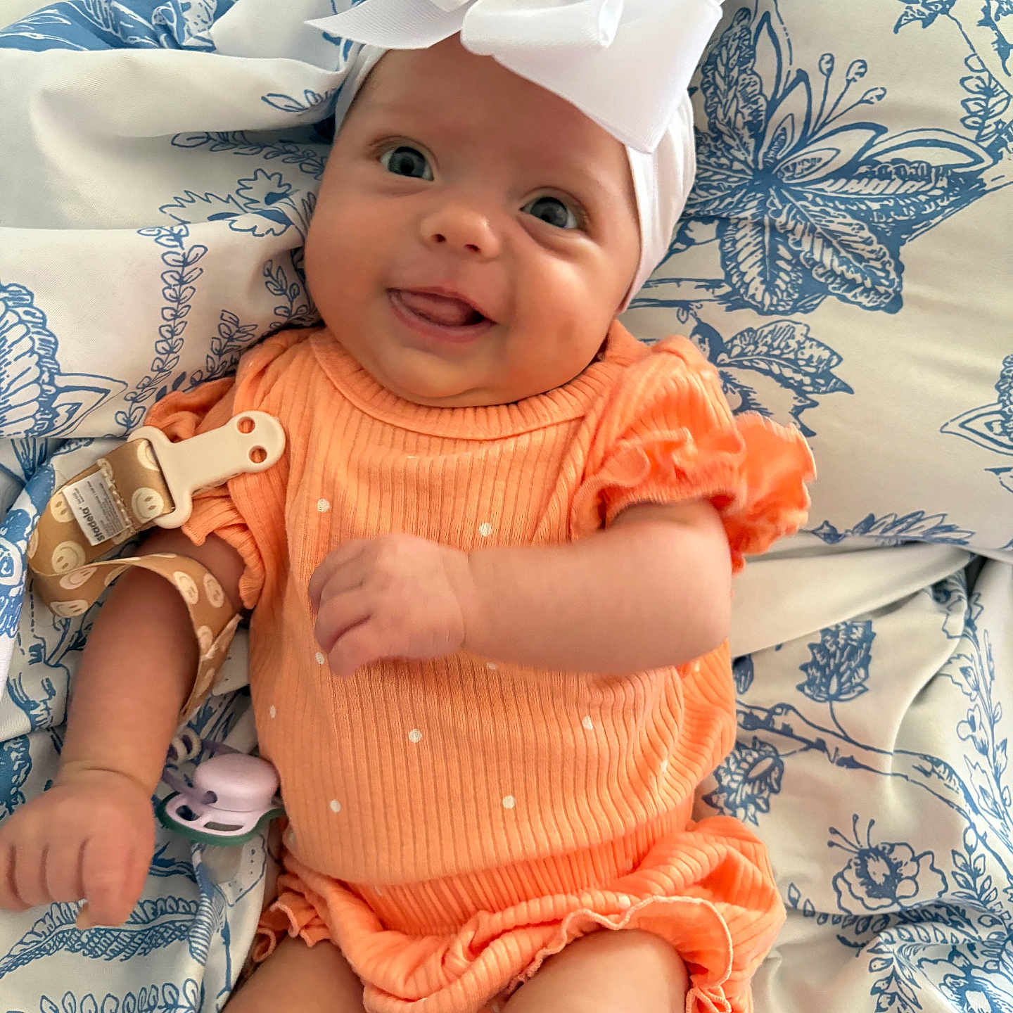 Violet is registered to the contest to win money with this photo: baby, blanket, blue, bow, child, clothing, cute, fabric, happy, headband, infant, lying_down, orange_outfit, pacifier, pattern, person, smiling, soft_toys, tongue_out, white