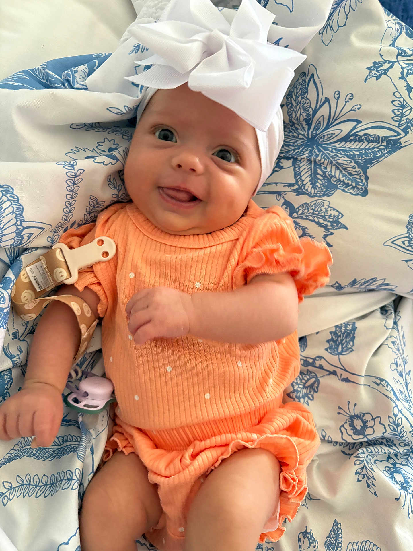 Violet is registered to the contest to win money with this photo: baby, infant, child, orange_outfit, headband, bow, blanket, pattern, blue, white, smiling, tongue_out, cute, happy, lying_down, fabric, clothing, soft_toys, pacifier, person