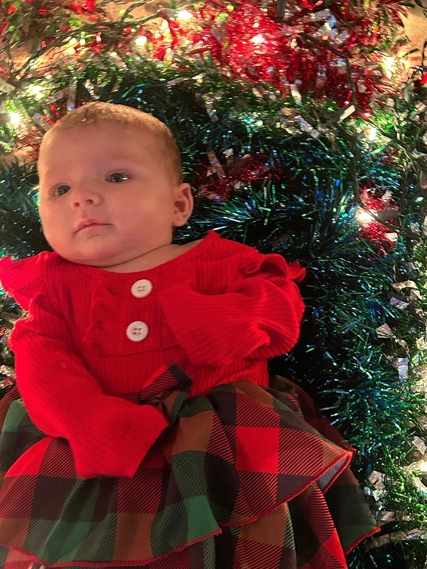 Phoebe is registered to the contest to win money with this photo: baby, infant, red_clothing, plaid_skirt, tinsel, holiday_lights, festive, christmas, sparkle, cute, face, portrait, child, young, indoors, celebration, colorful, decorations, smile, person