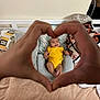 baby, child, hands, heart_shape, yellow_outfit, pillow, bed, smile, person, indoor, cozy, blanket, hand_gesture, cute, infant, cushion, happy, skin, family, love