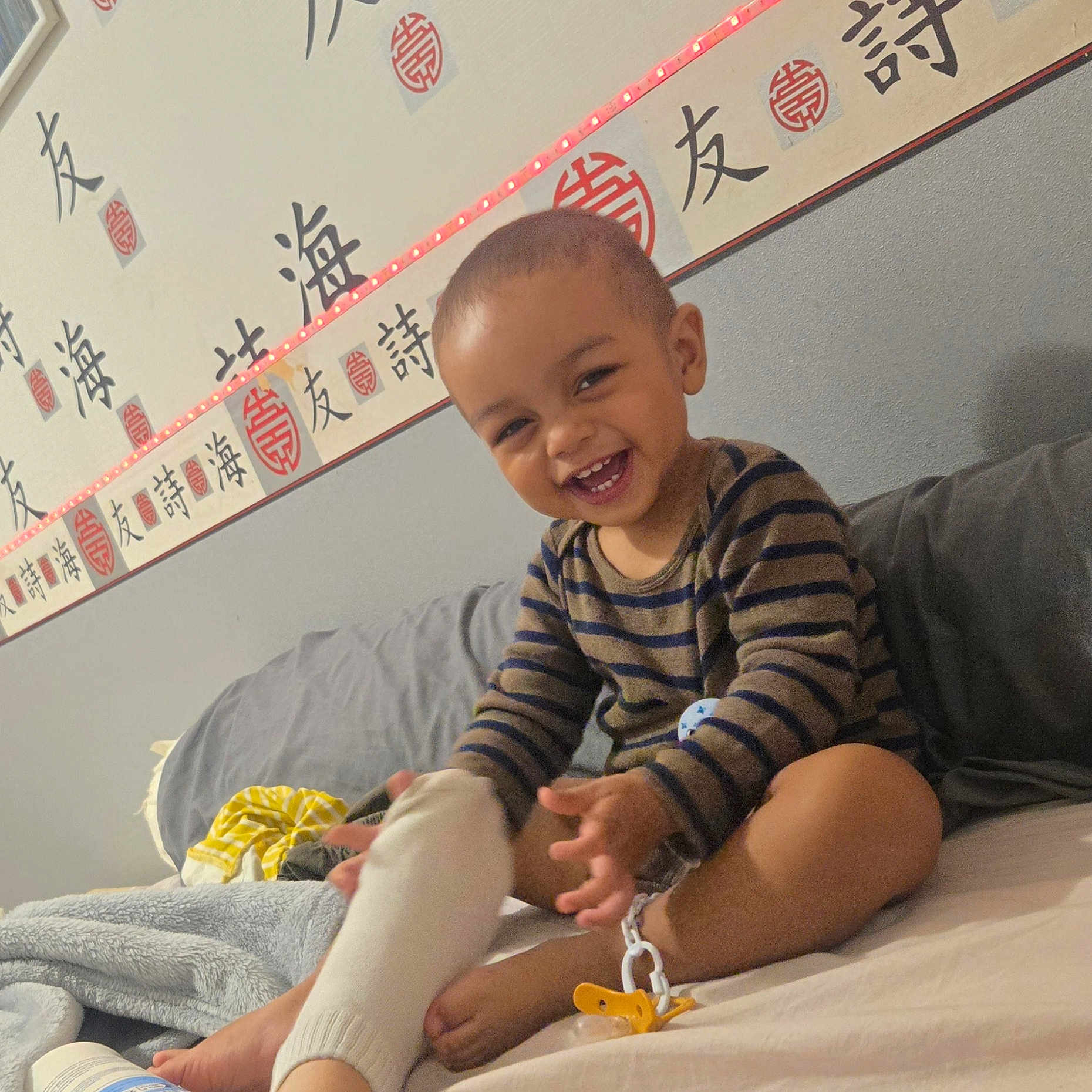 Mikalo a rejoint le concours — aidez-le/la à gagner de superbes lots ! asian_characters, bed, blanket, calligraphy, child, couch, foot, hand, happy, indoors, laughing, led_light, pajamas, person, playing, smiling, sock, striped_shirt, toddler, wall