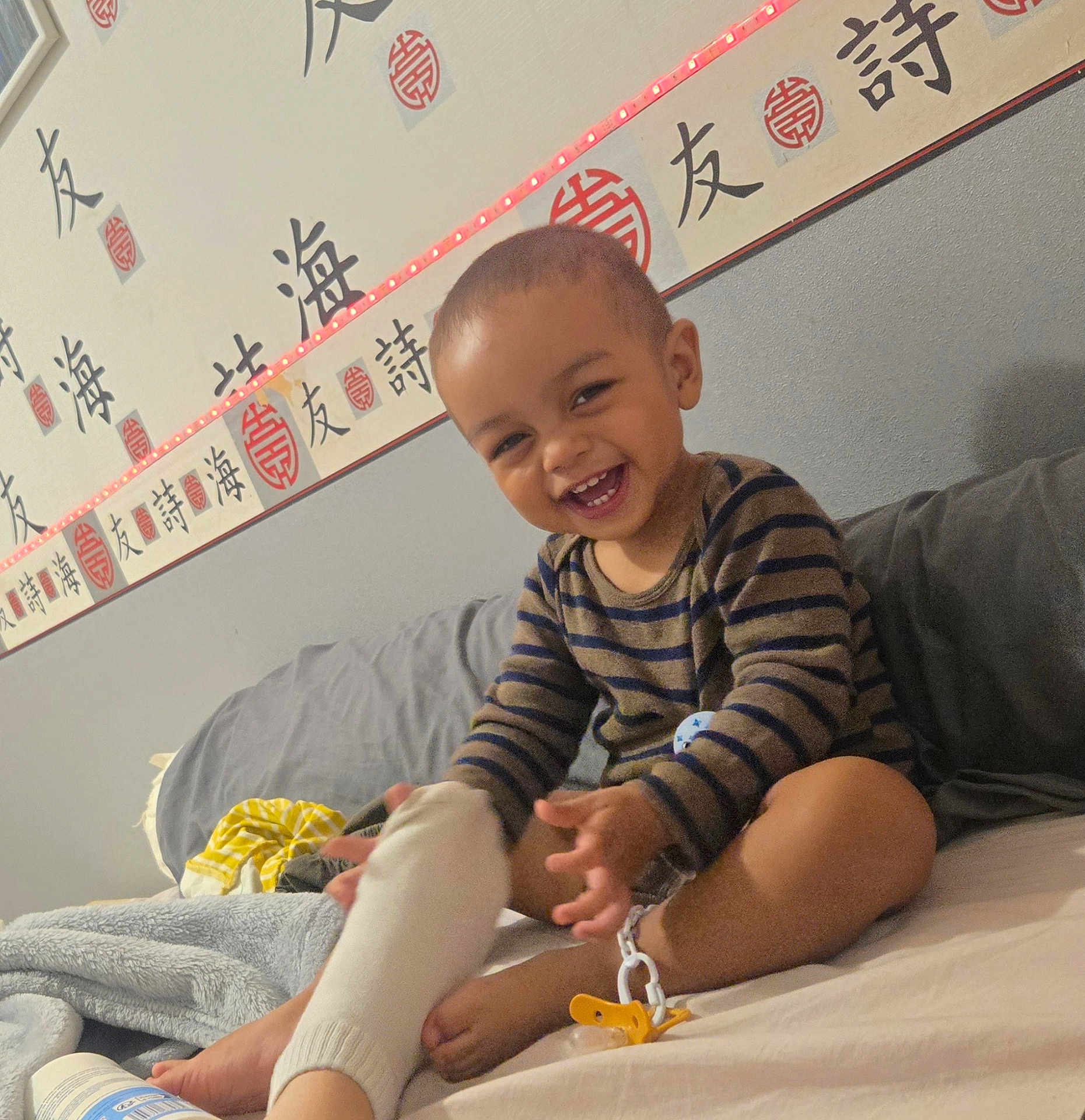 Mikalo a rejoint le concours — aidez-le/la à gagner de superbes lots ! toddler, child, smiling, laughing, striped_shirt, bed, sock, foot, hand, wall, calligraphy, asian_characters, led_light, pajamas, blanket, couch, indoors, happy, person, playing
