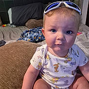 Asher is registered to the contest to win money with this photo: baby, child, infant, sitting, portrait, face, blue_eyes, blond_hair, sunglasses, pajamas, baby_clothing, smiling, indoor, bedroom, bed, blanket, pillow, couch, home, close_up