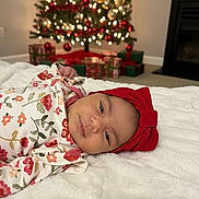 Daphne is registered to the contest to win money with this photo: baby, infant, red_bow, headband, floral_clothing, blanket, christmas_tree, decorations, presents, holiday, indoors, soft_texture, cute, smile, face, person, festive, warm, cozy, celebration