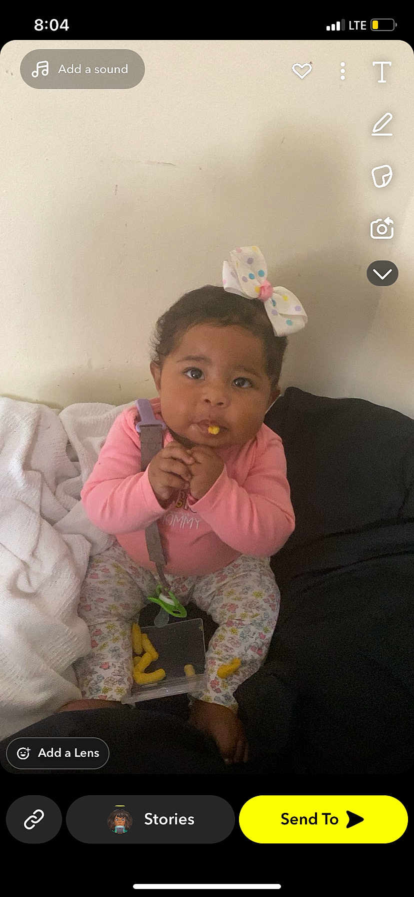 Kya is registered to the contest to win money with this photo: baby, child, cute, sitting, snack, cheese_puffs, bow, pink_clothing, floral_pants, blanket, couch, indoor, person, face, expression, food, playful, infant, home, cozy