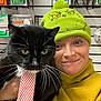 black_cat, cat, closeup, clothing, cute, face, green_hat, hat, holding, indoor, packaging, person, pet, portrait, shelf, smile, striped_tie, tie, whiskers, yellow_eyes