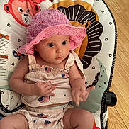 Azriella is registered to the contest to win money with this photo: baby, infant, pink_hat, american_flag, clothing, baby_seat, floor, wooden_floor, animal_pattern, monkey, lion, cute, child, indoor, person, seat, apparel, hat, sitting, curious