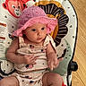 baby, infant, pink_hat, american_flag, clothing, baby_seat, floor, wooden_floor, animal_pattern, monkey, lion, cute, child, indoor, person, seat, apparel, hat, sitting, curious