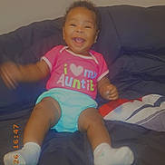 Serenity Brown is registered to the contest to win money with this photo: baby, child, smiling, sitting, couch, clothing, pink_shirt, socks, happy, indoor, face, person, cute, infant, joyful, home, pillow, arm_motion, text_on_clothing, portrait