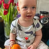 baby, blurry_background, casual_clothing, child, coffee_machine, cute, dinosaur_print, flower_vase, fruit, heart_emoji, indoor, kitchen, orange, person, portrait, red_bracelet, sitting, smiling, toddler, tulips