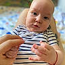 adult_hand, baby, blue_eyes, blurred_background, casual_clothing, child, close_up, cute, expression, fingers, hand, indoor, infant, intimate_moment, person, portrait, skin, soft_light, striped_shirt, young