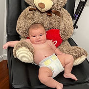 Azaiah is registered to the contest to win money with this photo: baby, diaper, teddy_bear, plush_toy, black_chair, heart, indoor, wooden_floor, relaxed, cute, child, infant, soft_toy, furniture, smiling, person, skin, comfort, seated, toy