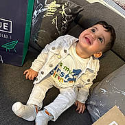Malek participe au concours pour gagner de l'argent avec cette photo : toddler, child, smile, sitting, white_jacket, colorful_shirt, socks, couch, pillow, shopping_bag, indoor, happy, face, head, person, home, celebration, birthday, clothing, cute