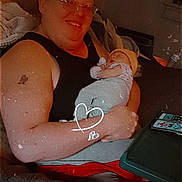 Jeremiah is registered to the contest to win money with this photo: adult, baby, smile, glasses, tattoo, sleeping, arm, clothing, blanket, chair, table, phone, hat, indoor, person, hand, relaxation, cozy, warm_lighting, heart_drawing