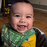 Mateo is registered to the contest to win money with this photo: baby, smiling, face, dimples, indoor, child, clothing, bib, yellow_shirt, happy, portrait, person, cheerful, young_child, cute, closeup, seat, wooden_floor, background_blur, expression