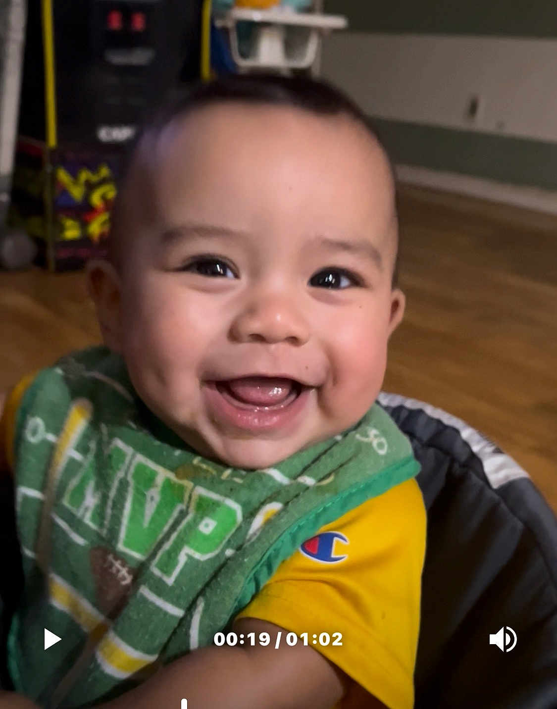 Mateo is registered to the contest to win money with this photo: baby, smiling, face, dimples, indoor, child, clothing, bib, yellow_shirt, happy, portrait, person, cheerful, young_child, cute, closeup, seat, wooden_floor, background_blur, expression