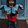 Chase is registered to the contest to win money with this photo: child, smile, helmet, shoulder_pads, football_gear, red_shirt, blue_jeans, sneakers, standing, door, doormat, outdoor, playful, protective_gear, fun, casual_clothing, young_boy, portrait, front_view, happy