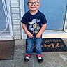 child, boy, glasses, smiling, standing, blue_door, transformers_shirt, jeans, sneakers, chalk_drawing, sidewalk, door_mat, hair_tied_up, outdoor, happy, concrete, front_door, casual_clothing, portrait, daylight