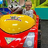 child, glasses, smile, indoor, play_area, arcade, ride, car, toy, red, yellow, cartoon_mouse, amusement, fun, entertainment, clothing, person, happy, playground, kid