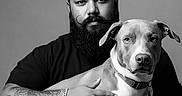 Atlas is registered to the contest to win money with this photo: man, beard, curly_hair, tattoo, bracelet, dog, pet, black_tshirt, jeans, portrait, studio, monochrome, grayscale, seated, calm, looking_at_camera, indoor, animal, friendship, companion