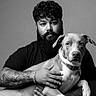 man, beard, curly_hair, tattoo, bracelet, dog, pet, black_tshirt, jeans, portrait, studio, monochrome, grayscale, seated, calm, looking_at_camera, indoor, animal, friendship, companion