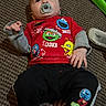 baby, child, pacifier, red_shirt, sesame_street, carpet, indoor, toy, slipper, pants, patches, cute, infant, lying_down, clothing, hand, foot, floor, person, casual
