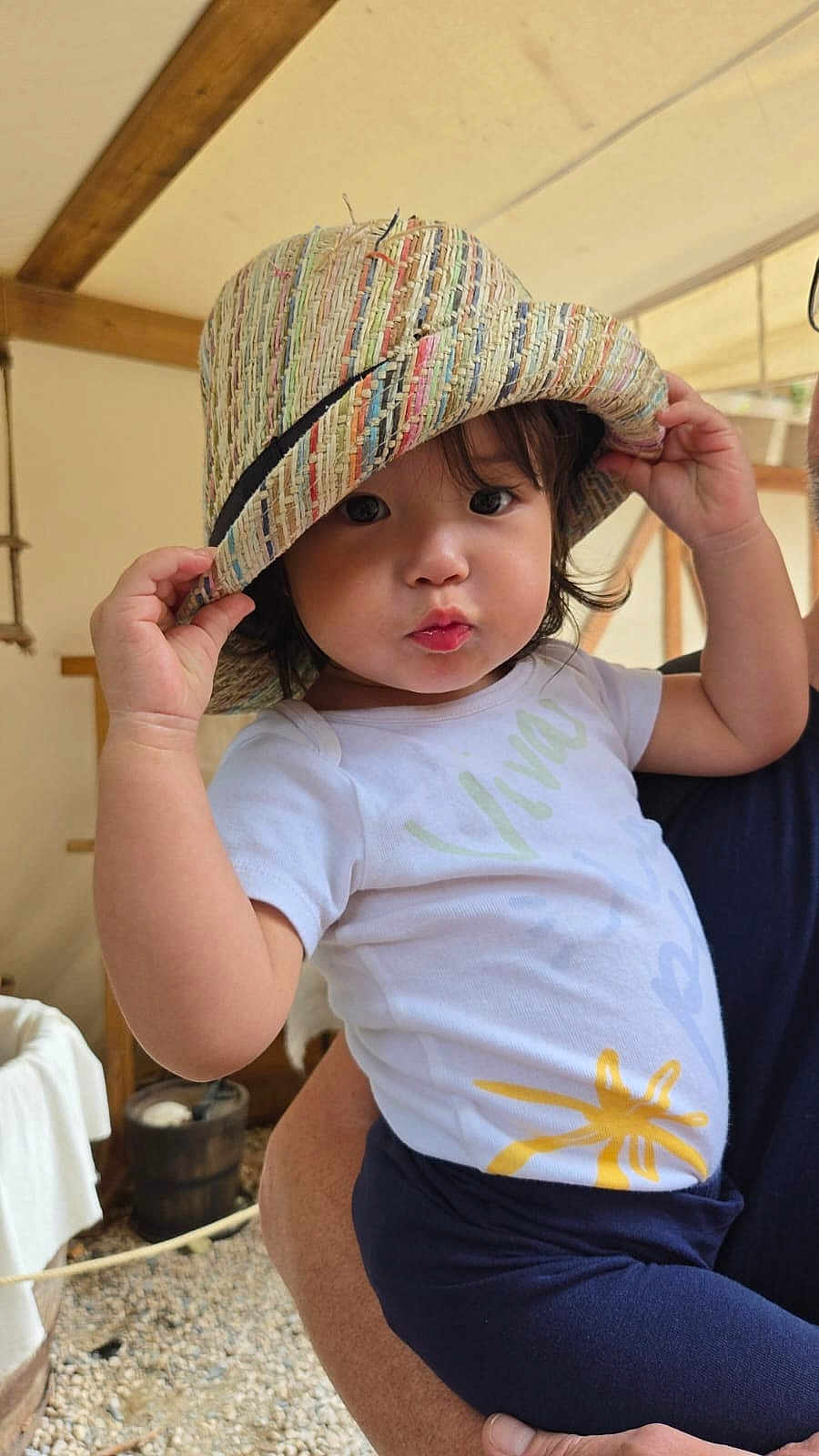 Olivia a rejoint le concours — aidez-le/la à gagner de superbes lots ! toddler, child, hat, straw_hat, indoor, person, holding, cute, face, eyes, curly_hair, white_shirt, navy_pants, hand, adult_arm, wooden_beams, background, natural_light, expression, playful