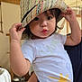 Olivia participe au concours pour gagner de l'argent avec cette photo : toddler, child, hat, straw_hat, indoor, person, holding, cute, face, eyes, curly_hair, white_shirt, navy_pants, hand, adult_arm, wooden_beams, background, natural_light, expression, playful