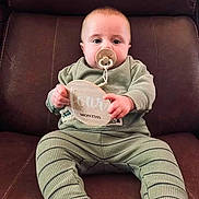 John is registered to the contest to win money with this photo: baby, child, infant, sitting, leather_chair, pacifier, milestone_sign, green_outfit, striped_clothing, bare_feet, portrait, indoor, furniture, couch, happy, wide_eyed, front_facing, close_up, family_photo, cute