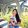 Camilo is registered to the contest to win money with this photo: baby, bench, blue_clothing, cap, child, daylight, garden, happy, hat, holding, long_hair, nature, outdoor, pergola, plants, portrait, smiling, sunlight, woman, wooden_bench